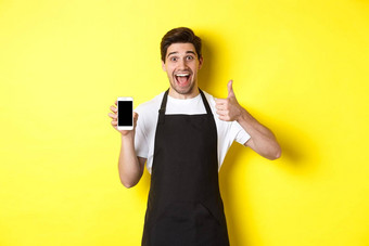 快乐咖啡师黑色的围裙显示智能<strong>手机屏</strong>幕使翘拇指推荐咖啡馆应用程序站黄色的背景