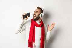 冬天假期技术概念男人。玩卡拉ok应用程序移动电话唱歌智能手机穿耳机站白色背景