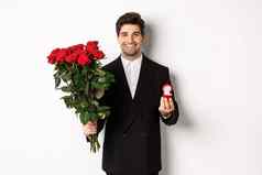 英俊的微笑男人。黑色的西装持有玫瑰订婚环使建议结婚站白色背景