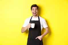 微笑年轻的咖啡师黑色的围裙持有咖啡杯站黄色的背景