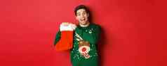 图像有吸引力的快乐的男人。毛衣持有圣诞节长袜礼物庆祝冬天假期站红色的背景