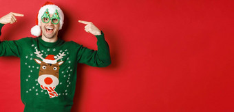 图像<strong>兴奋</strong>微笑的家伙指出<strong>聚会</strong>，派对眼镜庆祝一年站快乐的绿色毛衣圣诞老人他红色的背景