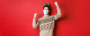 概念冠状病毒冬天假期社会距离图像兴奋快乐男人。医疗面具圣诞节服装提高手庆祝一年红色的背景