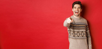 概念圣诞节庆祝活动冬天生活方式快乐有吸引力的男人。圣诞节毛衣祝快乐假期指出相机站红色的背景