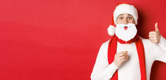 概念圣诞节冬天假期庆祝活动肖像有趣的男人。圣诞老人他持有胡子面具显示翘拇指享受一年聚会，派对站红色的背景