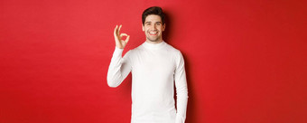 概念冬天假期圣诞节生活方式快乐英俊的男人。白色毛衣显示标志微笑很高兴推荐商店站红色的背景