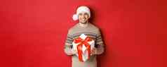 图像英俊的有胡子的男人。冬天毛衣圣诞老人他持有圣诞节礼物微笑现在包装丝带站红色的背景