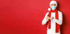 概念科维德圣诞节假期流感大流行快乐年轻的男人。圣诞老人他医疗面具清洁手防腐剂微笑站红色的背景