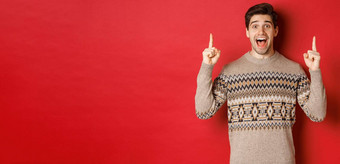 图像英俊的兴奋男人。感觉快乐圣诞节假期指出手指快乐的显示广告站毛衣红色的背景