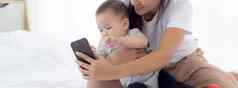 年轻的亚洲妈妈。婴儿女孩新生儿自拍聪明的电话床上卧室幸福妈妈女儿电话视频调用首页人家庭沟通概念