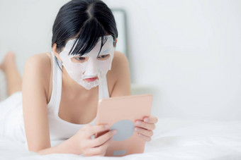 美丽的年轻的亚洲女人表面部面具说谎看平板电脑电脑床上卧室美女孩应用脸面具护肤品皱纹首页皮肤护理健康健康