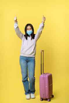 完整的长度拍摄快乐亚洲旅游女孩假期医疗脸面具显示拇指站手提箱黄色的背景