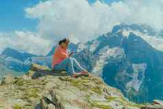女旅行者智能手机山完整的身体女人坐着岩石浏览智能手机多云的天空阳光明媚的一天山