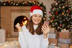 照片快乐微笑女孩读取x-mas新闻智能手机提出了的拳头坐着房子圣诞节装饰壁炉背景夫人穿圣诞老人他毛衣