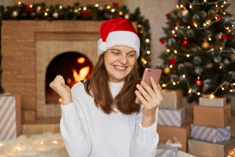 照片快乐微笑女孩读取x-mas新闻智能<strong>手机</strong>提出了的拳头坐着房子圣诞<strong>节</strong>装饰壁炉背景夫人穿圣诞老人他毛衣