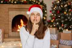女人圣诞老人他使安静的手势直接相机保持手指嘴唇女孩直头发摆姿势生活房间x-mas树壁炉背景
