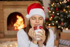 女人坐着装饰生活房间圣诞节树壁炉背景持有杯手享受热饮料眼睛关闭穿圣诞老人他