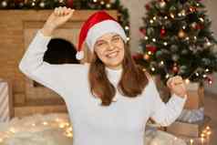 圣诞节夏娃快乐女人手跳舞愉快地相机露出牙齿的微笑夫人穿毛衣红色的圣诞老人他摆姿势生活房间装饰加兰x-mas树