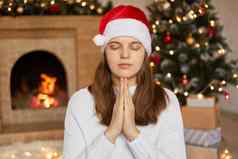 平静女穿圣诞老人老人他白色毛衣坐着室内圣诞节树壁炉保持手掌祈祷一年眼睛关闭
