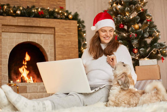 迷人的女人坐着地板上移动PC宠物坐在快乐女穿红色的他休闲毛衣夫人北京人的狗构成节日圣诞节房间壁炉