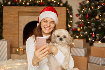 女孩使自拍不错的小狗坐着<strong>节日</strong>房间壁炉x-mas树女孩移动手机的屏幕迷人的微笑穿圣诞老人他休闲跳投
