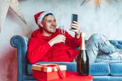 年轻的英俊的男人。圣诞老人老人他玻璃香槟进行生活广播朋友智能手机快乐的家伙庆祝圣诞节在线互联网说谎沙发上首页