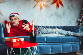 年轻的英俊的男人。圣诞老人老人他玻璃香槟采取自拍智能手机有趣的的家伙拍摄移动电话说谎沙发上概念圣诞节庆祝<strong>活动首页</strong>