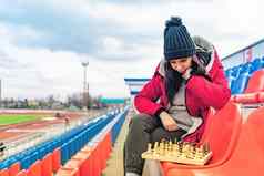 年轻的女人冬天衣服戏剧国际象棋坐着体育场看台女黑色的帽国际象棋体育体育场多云的天气