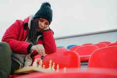 年轻的女人冬天衣服戏剧国际象棋坐着体育场看台女黑色的帽国际象棋体育体育场多云的天气