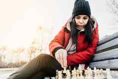 年轻的女人冬天衣服戏剧国际象棋坐着板凳上城市公园女黑色的帽国际象棋木皮尤多云的天气