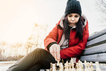 年轻的女人冬天衣服戏剧国际象棋坐着板凳上城市公园女黑色的帽国际象棋木皮尤多云的天气