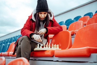 年轻的女人冬天衣服戏剧国际象棋坐着<strong>体育场</strong>看台女黑色的帽国际象棋体育<strong>体育场</strong>多云的天气