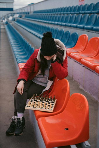 年轻的女人冬天衣服戏剧国际象棋坐着<strong>体育场</strong>看台女黑色的帽国际象棋体育<strong>体育场</strong>多云的天气