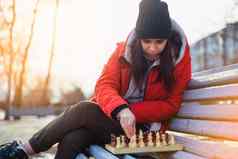 年轻的女人冬天衣服戏剧国际象棋坐着板凳上城市公园女黑色的帽国际象棋木皮尤多云的天气