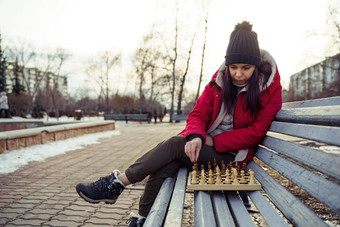 年轻的女人冬天衣服戏剧国际象棋坐着板凳上城市公园女黑色的帽国际象棋木皮尤多云的天气