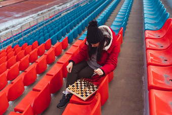 年轻的女人冬天衣服戏剧国际象棋坐着<strong>体育</strong>场看台女黑色的帽国际象棋<strong>体育体育</strong>场多云的天气