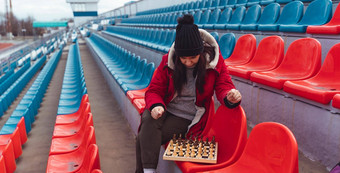 年轻的女人冬天衣服戏剧国际象棋坐着<strong>体育场</strong>看台女黑色的帽国际象棋体育<strong>体育场</strong>多云的天气