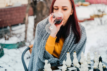 年轻的女人玩国际象棋喝红色的酒院子里女包装灰色格子酒精坐着街玩董事会游戏冬天季节