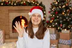 年轻的女人微笑幸福的高高兴兴地挥舞着手欢迎问候再见穿圣诞老人他毛衣摆姿势圣诞节树壁炉盒子礼物