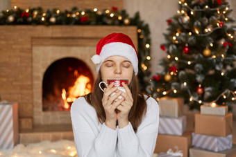 圣诞节女人喝热茶咖啡享受饮料关闭眼睛持有杯手坐着生活房间圣诞节装饰女孩温暖的毛衣圣诞老人他