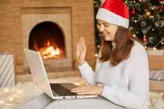 美丽的年轻的女人圣诞老人他问候圣诞节一年在线坐着移动PC膝盖背景圣诞节树壁炉挥舞着手相机
