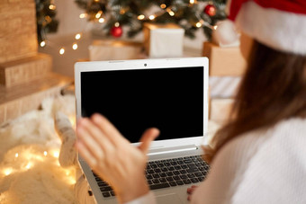 照片迷人的夫人坐着持有移动PC挥舞着棕榈屏幕穿圣诞老人他白色毛衣装饰x-mas生活房间向后视图女视频调用