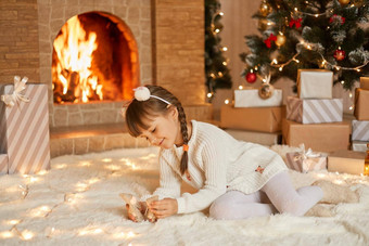 孩子玩玩具圣诞节早....坐着地毯装饰生活房间x-mas树壁炉微笑快乐女孩猪尾持有小玩具手一年