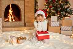 迷人的女孩坐着地板上现在盒子保持手掌脸颊无聊等待开放礼物摆姿势圣诞节树壁炉背景穿圣诞老人他