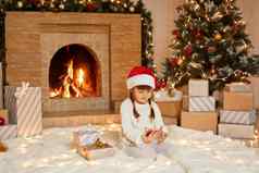可爱的女孩电话手坐着生活房间地板上礼服节日他休闲服装玩游戏智能手机圣诞节夏娃摆姿势圣诞节树壁炉软地毯