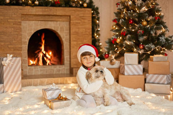 女孩狗圣诞节夏娃坐着白色软地毯孩子相机拥抱宠物女孩子圣诞老人他坐着火的地方冷杉树