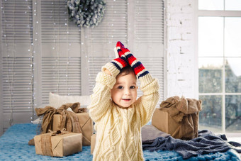有趣的好玩的男孩孩子坐在床上圣诞节一天礼物盒子白色羊毛针织毛衣大明亮的连指手套笑着说大声室内节日装饰