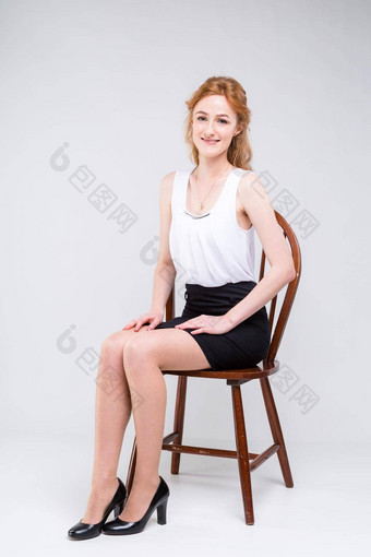 年轻的美丽的女人长红色的卷曲的头发坐着木椅子<strong>白色</strong>背景工作室穿着<strong>白色</strong>上衣<strong>短袖</strong>黑色的短裙子高高跟鞋