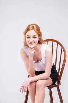 年轻的美丽的女人长红色的卷曲的头发坐着木椅子白色背景工作室穿着白色上衣短袖黑色的短裙子高高跟鞋