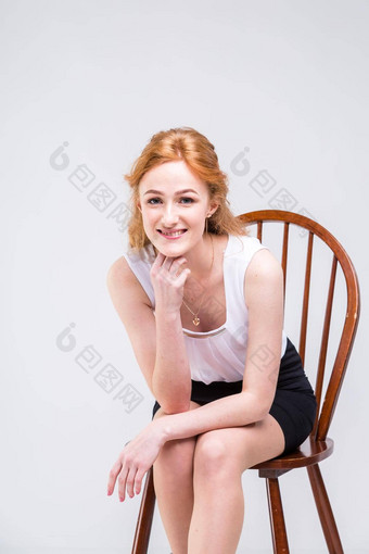 年轻的美丽的女人长红色的卷曲的头发坐着木椅子白色背景工作室穿着白色上衣短袖黑色的短裙子高高跟鞋